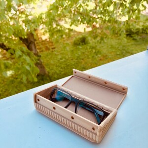 May include: A beige, rectangular glasses case with a textured design and a pair of black glasses inside.
