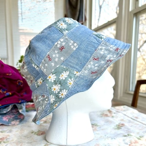 May include: A blue denim bucket hat with a patchwork design featuring white daisies and red ribbon accents.