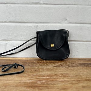 May include: A small, black leather crossbody bag with a flap closure and a gold-colored button. The bag has a long, thin strap and is set against a white brick wall and a wooden surface.