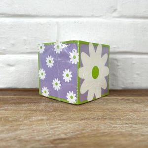 May include: A square, decorative block with a floral design. The block features a light purple background with white daisy-like flowers. One side has a large white flower with a green center. The edges are outlined in green.