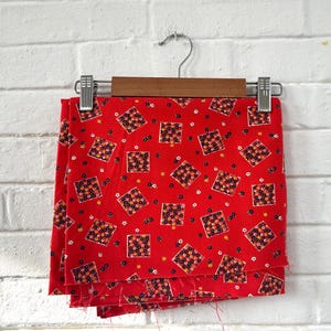 May include: Red fabric with a repeating pattern of small floral squares and tiny white and yellow flowers. The fabric is hanging on a wooden hanger with a metal clip. The fabric is likely vintage.