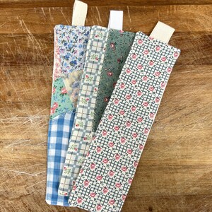 May include: Four fabric bookmarks with floral and gingham patterns. Each bookmark has a different design, including blue and white gingham, small pink flowers on a white grid, and a patchwork design with floral and solid fabrics. Each has a cream-colored ribbon.