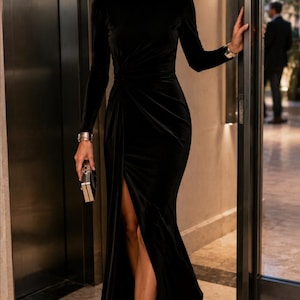 May include: A floor-length black velvet gown featuring long sleeves, a high neckline, and a gathered waist detail that creates a side slit. The dress is paired with black pointed-toe heels, a small metallic clutch, and large silver statement earrings.