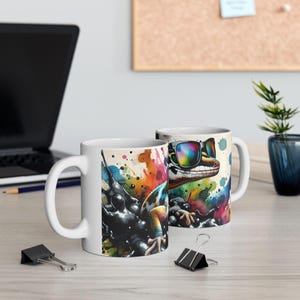 Peut inclure: Deux mugs en céramique blanche avec un motif de gecko coloré. L'un des mugs présente un gecko avec des lunettes de soleil, l'autre un gecko avec des éclaboussures de peinture. Les mugs ont une anse blanche et sont sur une surface en bois.