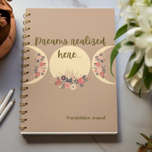 Könnte beinhalten: Ein braunes Manifestationsjournal mit goldener Spiralbindung. Der Einband zeigt ein florales Design mit drei Mondsicheln und dem Text "Dreams realized here..." in goldenen Lettern. Am unteren Rand des Einbands steht "Manifestation Journal".
