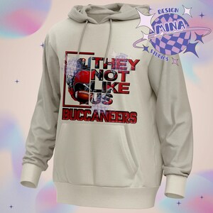 Pu&ograve; includere: Una felpa con cappuccio grigia chiaro con una stampa grafica rossa e nera di un casco da football e il testo "They Not Like Us Buccaneers".