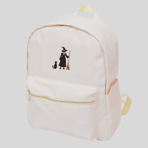 May include: A pink canvas backpack with a black embroidered image of a witch holding a broom and a black cat walking beside her.