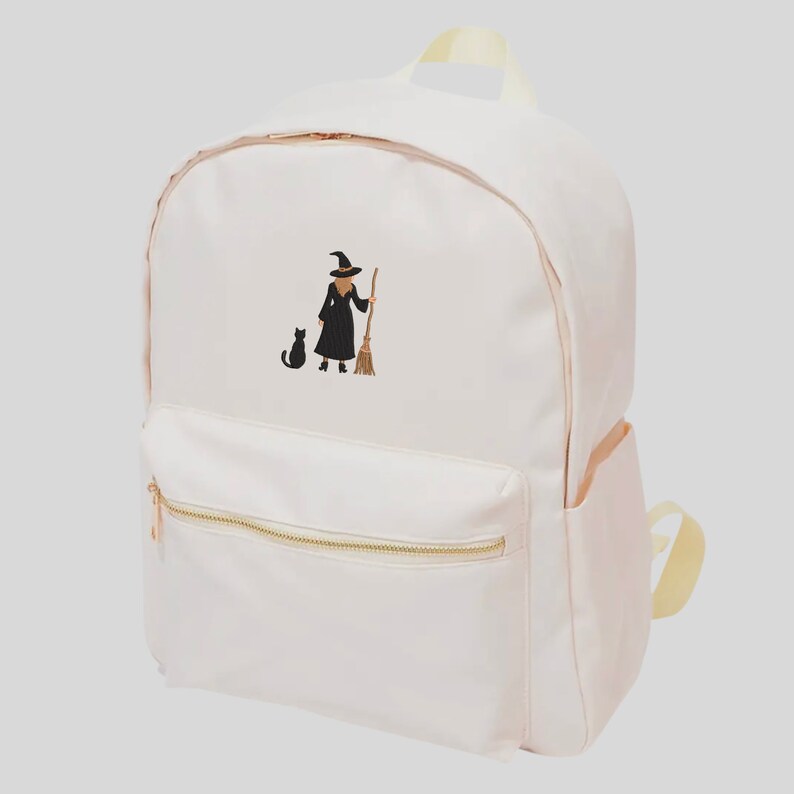 May include: A pink canvas backpack with a black embroidered image of a witch holding a broom and a black cat walking beside her.