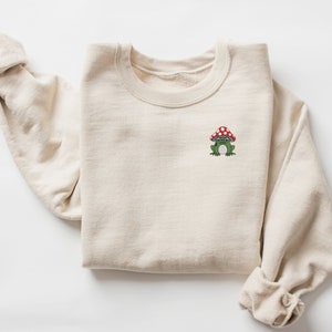 May include: A beige sweatshirt with a green frog wearing a red and white mushroom hat embroidered on the front.