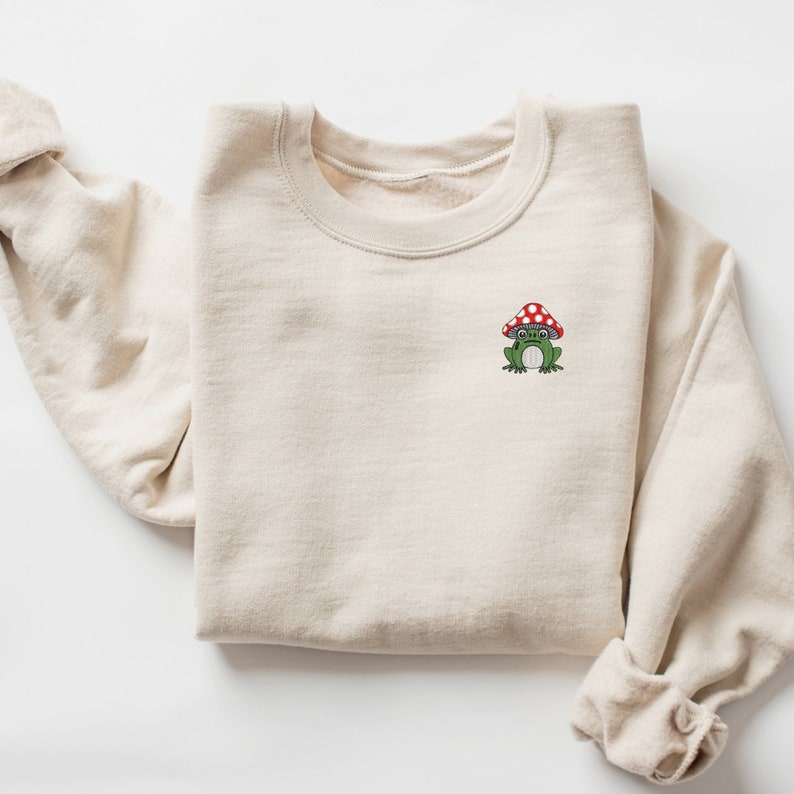 May include: A beige sweatshirt with a green frog wearing a red and white mushroom hat embroidered on the front.