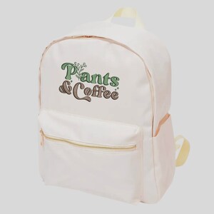 May include: A cream-colored backpack with a front pocket and gold-colored zippers. The backpack features the text "Plants & Coffee" in green and brown lettering with a botanical design. The bag has a top handle and adjustable straps.