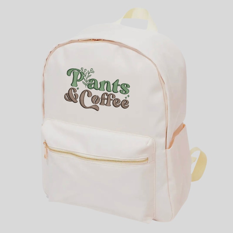 May include: A cream-colored backpack with a front pocket and gold-colored zippers. The backpack features the text "Plants & Coffee" in green and brown lettering with a botanical design. The bag has a top handle and adjustable straps.