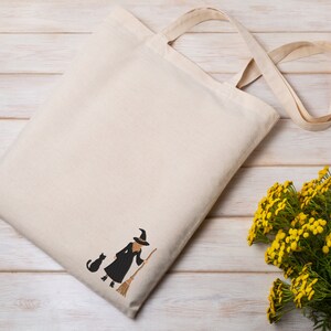 May include: A white tote bag with a black embroidered witch and cat design. The bag has long straps and is made of natural fabric.