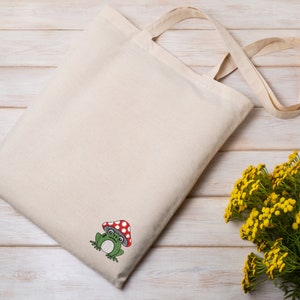 May include: A white tote bag with a green frog wearing a red and white mushroom hat embroidered on the front. The bag has long straps and is lying on a white wooden surface.