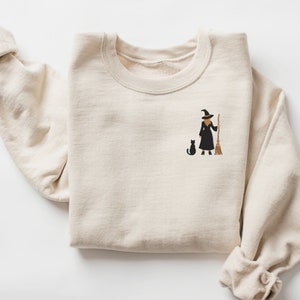 May include: A beige sweatshirt with an embroidered image of a witch holding a broom and a black cat. The witch is wearing a black dress and a pointed hat.
