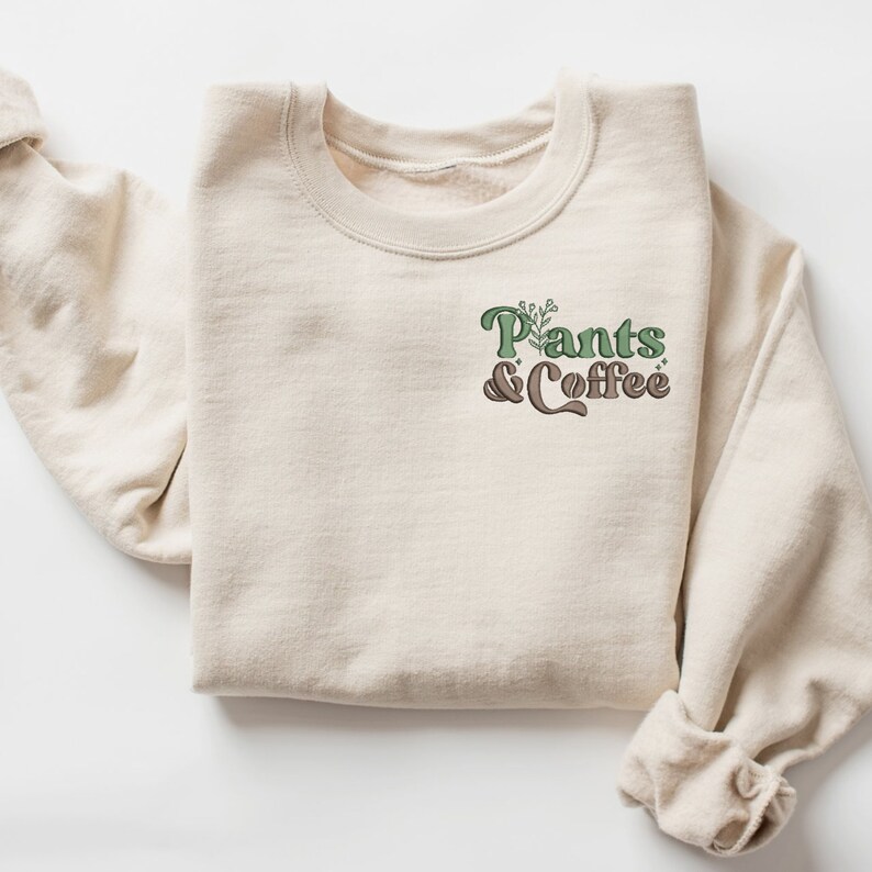 May include: A cream-colored sweatshirt with the text "Pants & Coffee" in green and brown. The design includes a plant graphic above the words. The sweatshirt is folded neatly, showcasing the design on the front.