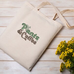 May include: A cream-colored tote bag with the words "Plants & Coffee" in green and brown lettering. The bag has long, natural-colored handles and is made of a sturdy fabric. A bouquet of yellow flowers sits to the side.