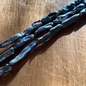 May include: Close-up of several strands of dark blue-grey baroque pearls. The pearls have an iridescent sheen, reflecting hints of green and purple. The pearls are strung together, creating a necklace or bracelet.