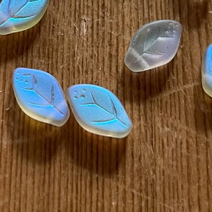 May include: Several iridescent leaf-shaped beads with a blue and yellow color gradient. The beads have a textured surface and a detailed leaf vein design. The beads are scattered on a wooden surface.