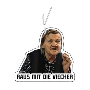 May include: A white car air freshener featuring a photograph of an older woman with a stern expression. The text "RAUS MIT DIE VIECHE" is printed in black at the bottom of the air freshener.