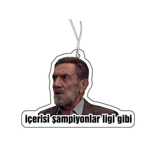 May include: Air freshener featuring a man's portrait with the text "içerisi şampiyonlar ligi'gibi". The air freshener is white with a black outline and a string for hanging. The man has a beard and is wearing a suit.