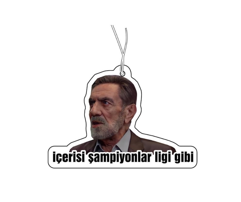 May include: Air freshener featuring a man's portrait with the text "i&ccedil;erisi şampiyonlar ligi'gibi". The air freshener is white with a black outline and a string for hanging. The man has a beard and is wearing a suit.