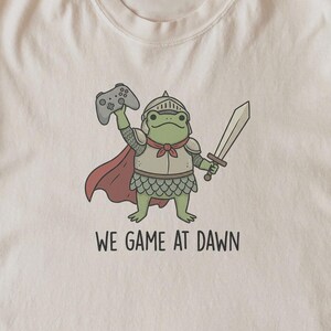 May include: A cream-colored t-shirt featuring a graphic of a green frog dressed in knight armor, a red cape, and a helmet. The frog holds a gray video game controller in one hand and a sword in the other. Below the graphic is the text WE GAME AT DAWN.