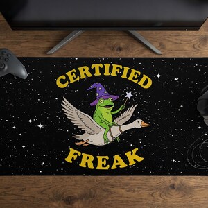 May include: A rectangular black desk mat featuring a starry night sky background. In the center, a green frog wearing a purple wizard hat and holding a star wand rides a white goose. The yellow text reads CERTIFIED FREAK in a bold font.