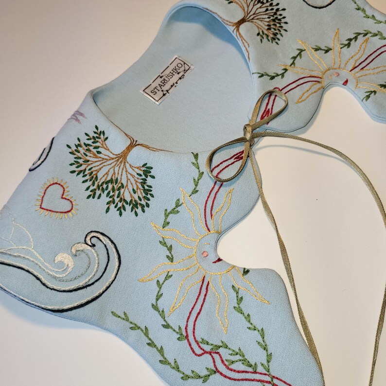 May include: A light blue embroidered collar with a whimsical design. The collar features embroidered trees, suns, hearts, and waves in shades of green, yellow, red, and black. The brand name "STARUSHKO" is visible on a tag.