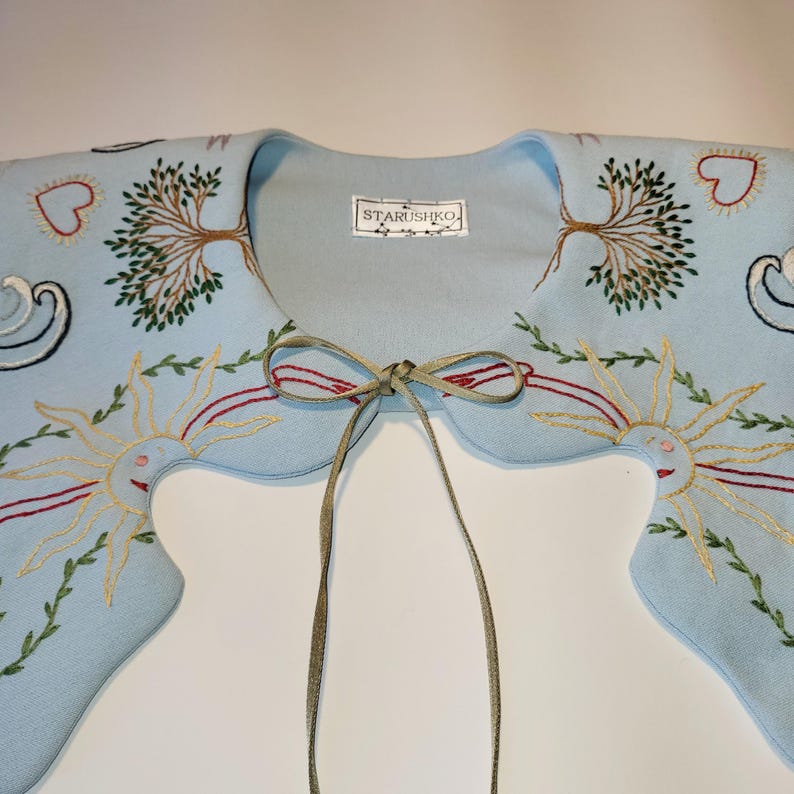 May include: A light blue embroidered collar with a tie closure. The collar features embroidered suns, trees, hearts, and swirls in shades of gold, red, and green. The label reads "STARUSHKO".