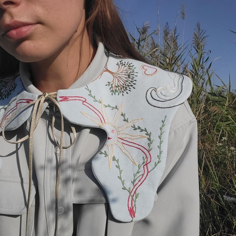 May include: A light blue embroidered collar with a sun, tree, and wave design. The collar is tied with a gold ribbon. The embroidery features red, green, and yellow thread. The collar is worn over a light gray button-up shirt.