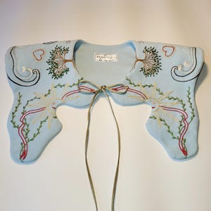 May include: A light blue embroidered collar with a suede tie. The collar features embroidered suns, trees, hearts, and swirling designs in red, green, and cream colors. The tag reads "STARUSHIKO".