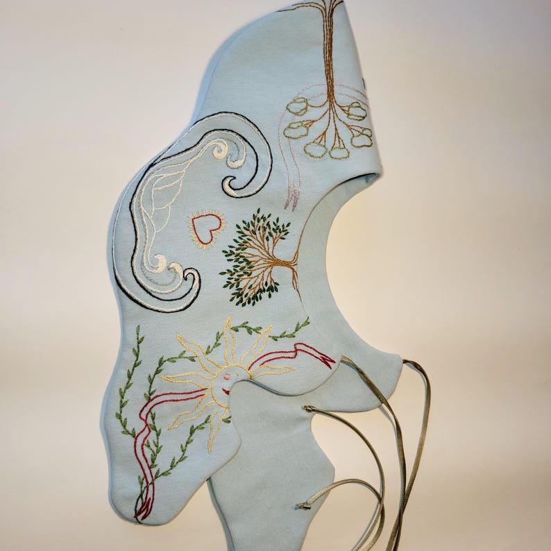 May include: A light blue, embroidered hood with a unique shape. The hood features embroidered designs including a tree, sun, and swirling patterns in various colors. The hood has a drawstring closure and is made of a soft material.