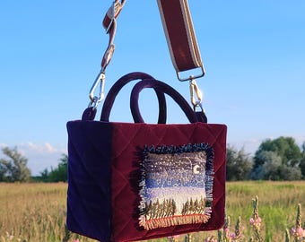 Puede incluir: Un pequeño bolso acolchado con un exterior de terciopelo morado oscuro. El bolso tiene un asa superior y una correa larga. La parte delantera del bolso presenta un parche bordado rectangular que representa un cielo nocturno con una luna creciente, estrellas y una fila de árboles.
