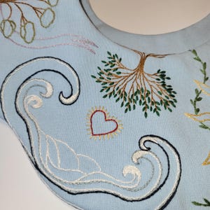 May include: Light blue fabric with embroidered designs. Features a tree, heart, and wave patterns in various colors. The embroidery includes white, black, red, yellow, and green threads, creating a detailed and artistic textile.