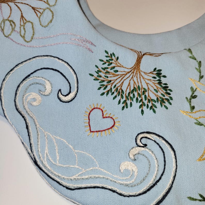 May include: Light blue fabric with embroidered designs. Features a tree, heart, and wave patterns in various colors. The embroidery includes white, black, red, yellow, and green threads, creating a detailed and artistic textile.