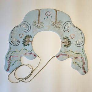 May include: A light blue, embroidered collar with a central opening. The collar features detailed embroidery of trees, hearts, suns, and swirling patterns in various colors. A thin, olive-green cord extends from the collar.