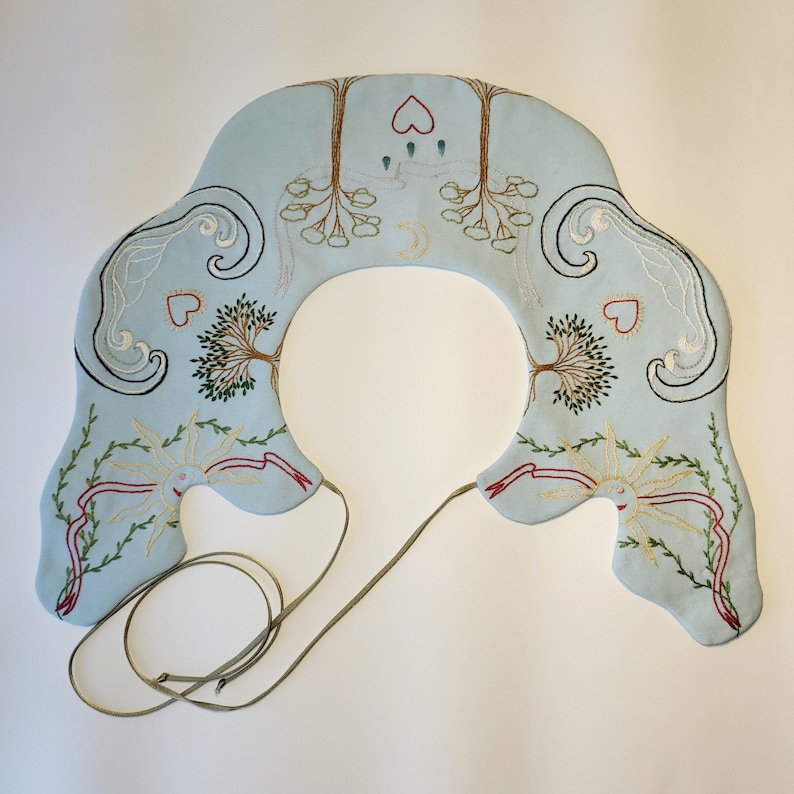 May include: A light blue, embroidered collar with a central opening. The collar features detailed embroidery of trees, hearts, suns, and swirling patterns in various colors. A thin, olive-green cord extends from the collar.