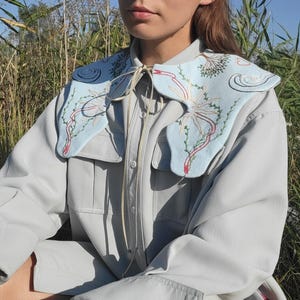 May include: A light blue collar with embroidered floral and abstract designs. The collar is worn over a light gray button-up shirt. The embroidery features red, green, and white accents. The collar is tied with a cream-colored string.