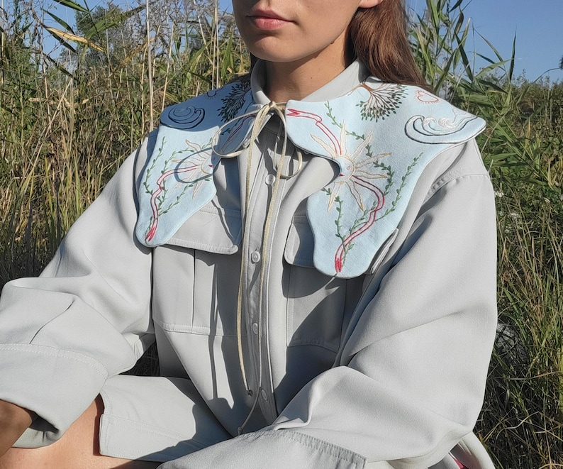 May include: A light blue collar with embroidered floral and abstract designs. The collar is worn over a light gray button-up shirt. The embroidery features red, green, and white accents. The collar is tied with a cream-colored string.