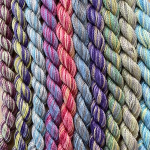 May include: Close-up of colourful embroidery floss in shades of purple, pink, blue, and yellow. The floss is arranged in diagonal rows, creating a textured display. Suitable for embroidery, cross-stitch, or other craft projects.