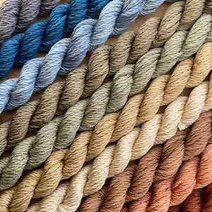 May include: A collection of 12 skeins of yarn in various shades of blue, green, brown, and beige. The yarn is arranged in a gradient from dark blue to light beige.