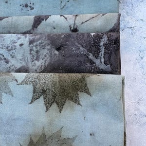 May include: A stack of three folded fabric pieces featuring botanical prints. The top piece is light blue with dark speckles, the middle is a dark charcoal grey with leaf patterns, and the bottom is light blue with dark green maple leaf prints.