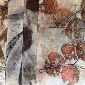May include: A collection of fabric swatches featuring botanical eco-prints. The textiles display various leaf patterns in shades of rust, brown, and muted grey on a light, off-white background, showcasing natural plant-based pigment transfers.