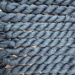 May include: Close-up of several twisted skeins of blue yarn. The yarn is a muted blue-grey colour, with a slightly textured appearance. The skeins are arranged in parallel rows, creating a pattern of thick, rope-like strands.