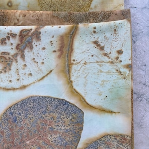 May include: A stack of folded fabric pieces featuring botanical prints. The textile displays natural leaf imprints in shades of brown, tan, and muted blue-grey against a light cream background, showcasing intricate vein patterns and organic textures.