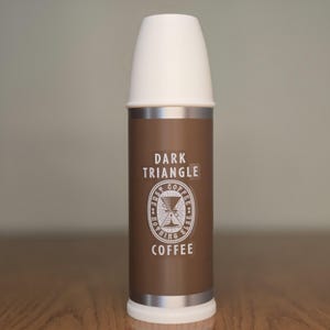 May include: A brown and silver coffee thermos with a white lid. The thermos has the words "DARK TRIANGLE COFFEE" printed on it, along with a logo. The thermos is cylindrical and appears to be made of metal.