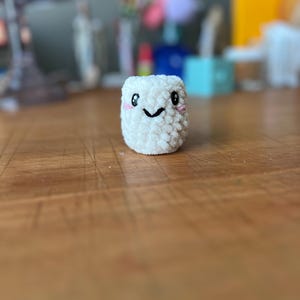 May include: A white crocheted plush toy shaped like a marshmallow with a smiling face and pink cheeks.