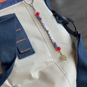 May include: A white and blue striped beaded keychain with the word "SAIL AWAY" spelled out in gold letters. The keychain has a red and white striped bead and a gold charm of a ship's wheel.