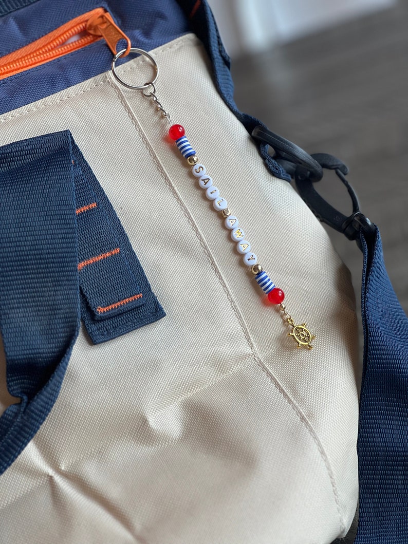 May include: A white and blue striped beaded keychain with the word "SAIL AWAY" spelled out in gold letters. The keychain has a red and white striped bead and a gold charm of a ship's wheel.